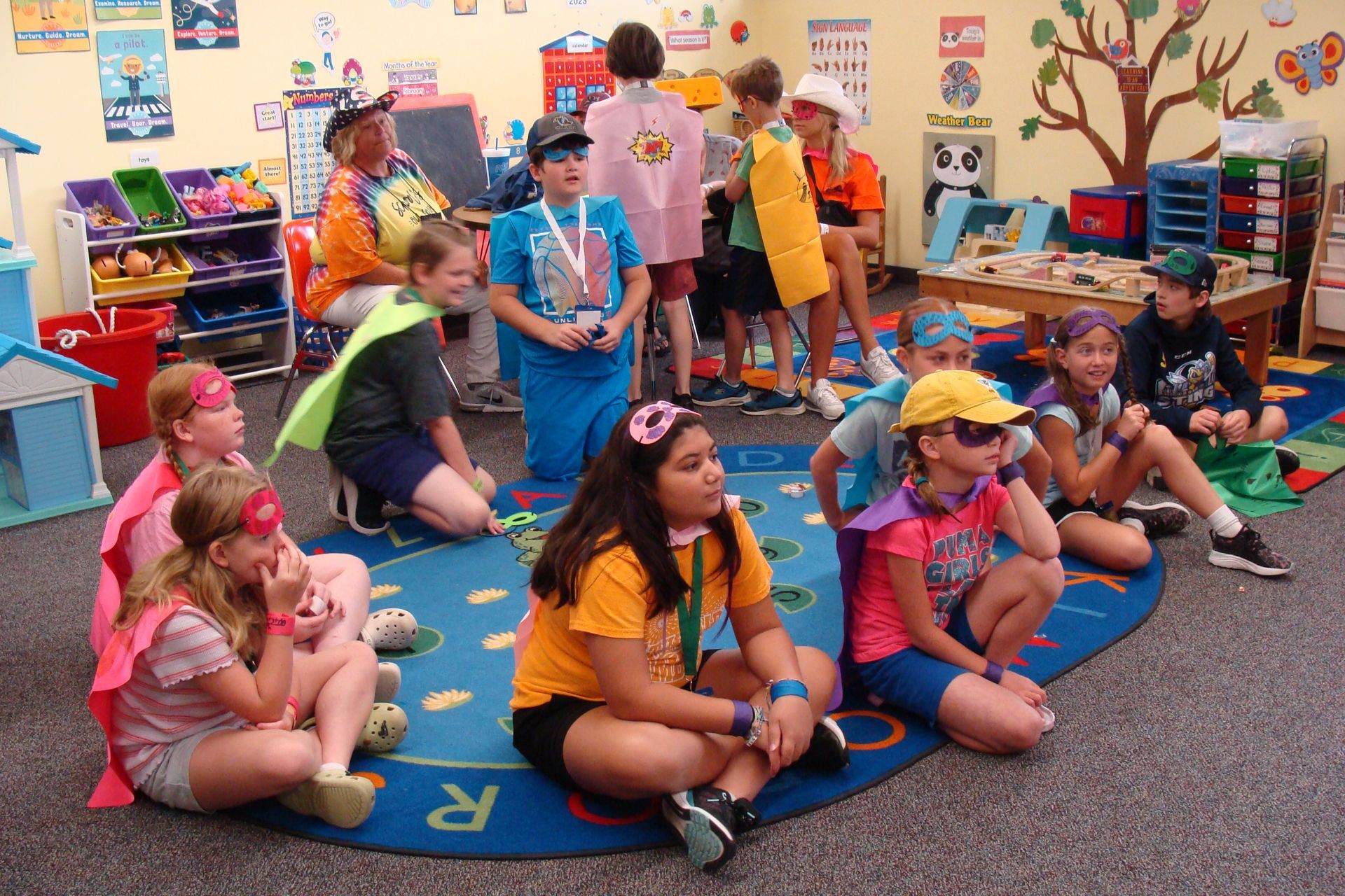 Children sitting on a classroom rug during a group activity, with toys and posters in the background.