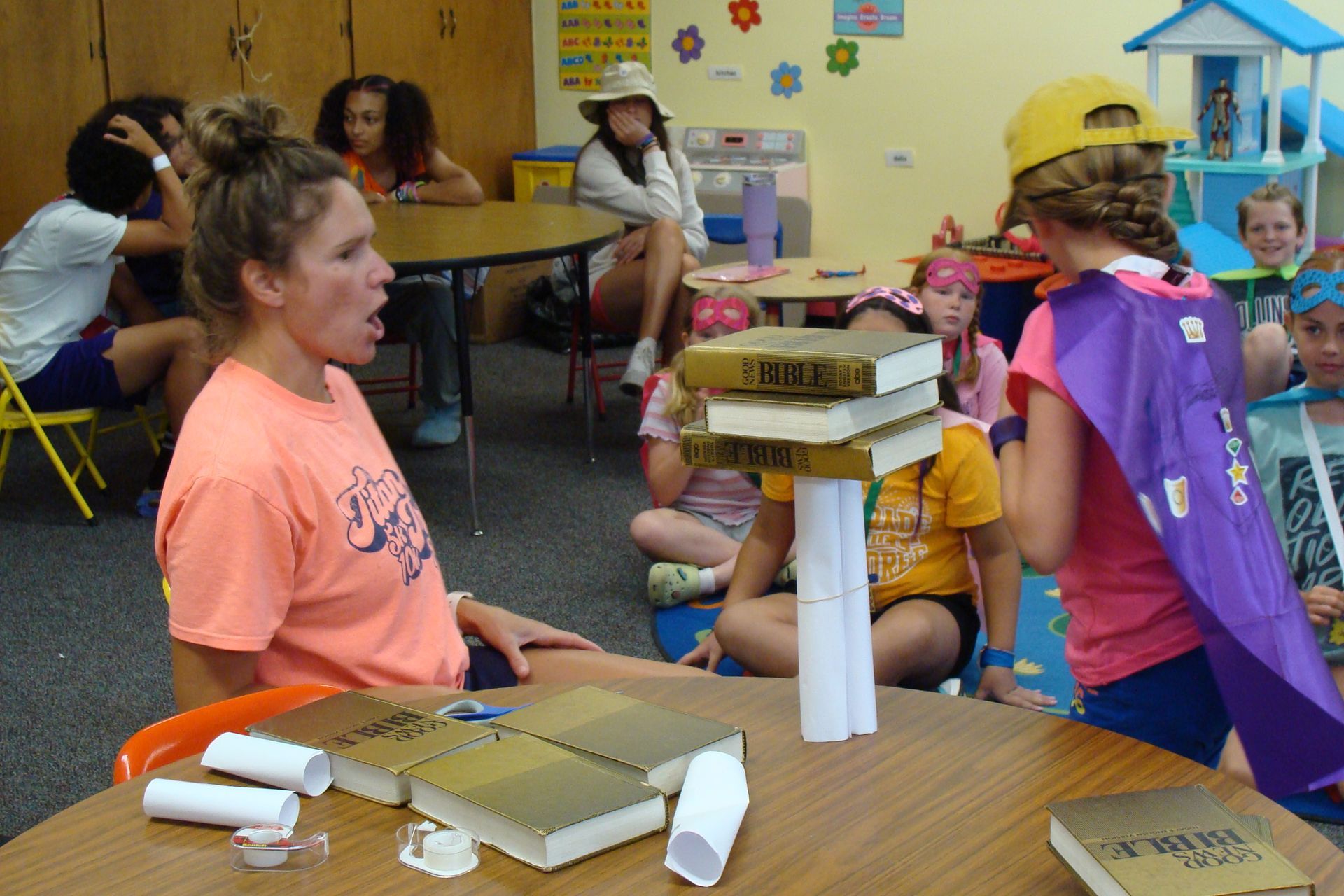 Children in a classroom wearing cardboard helmets and capes, gathered around tables and stacked books