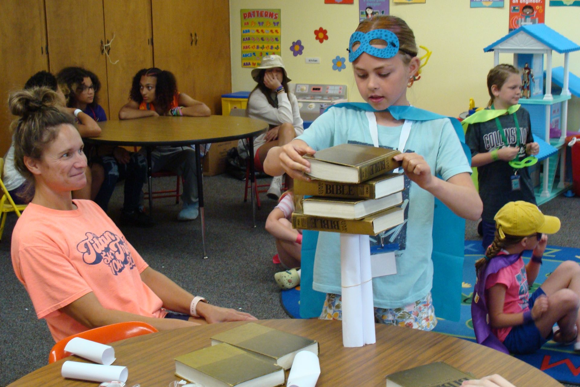 Child stacking books on a tower while seated adults watch in a classroom activity area