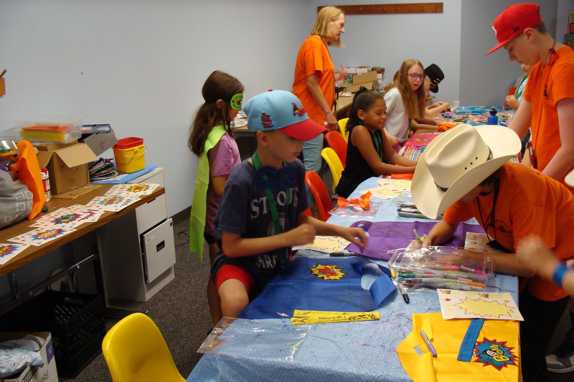 Children doing arts and crafts at tables in a classroom, with papers, crayons, and supplies scattered around.