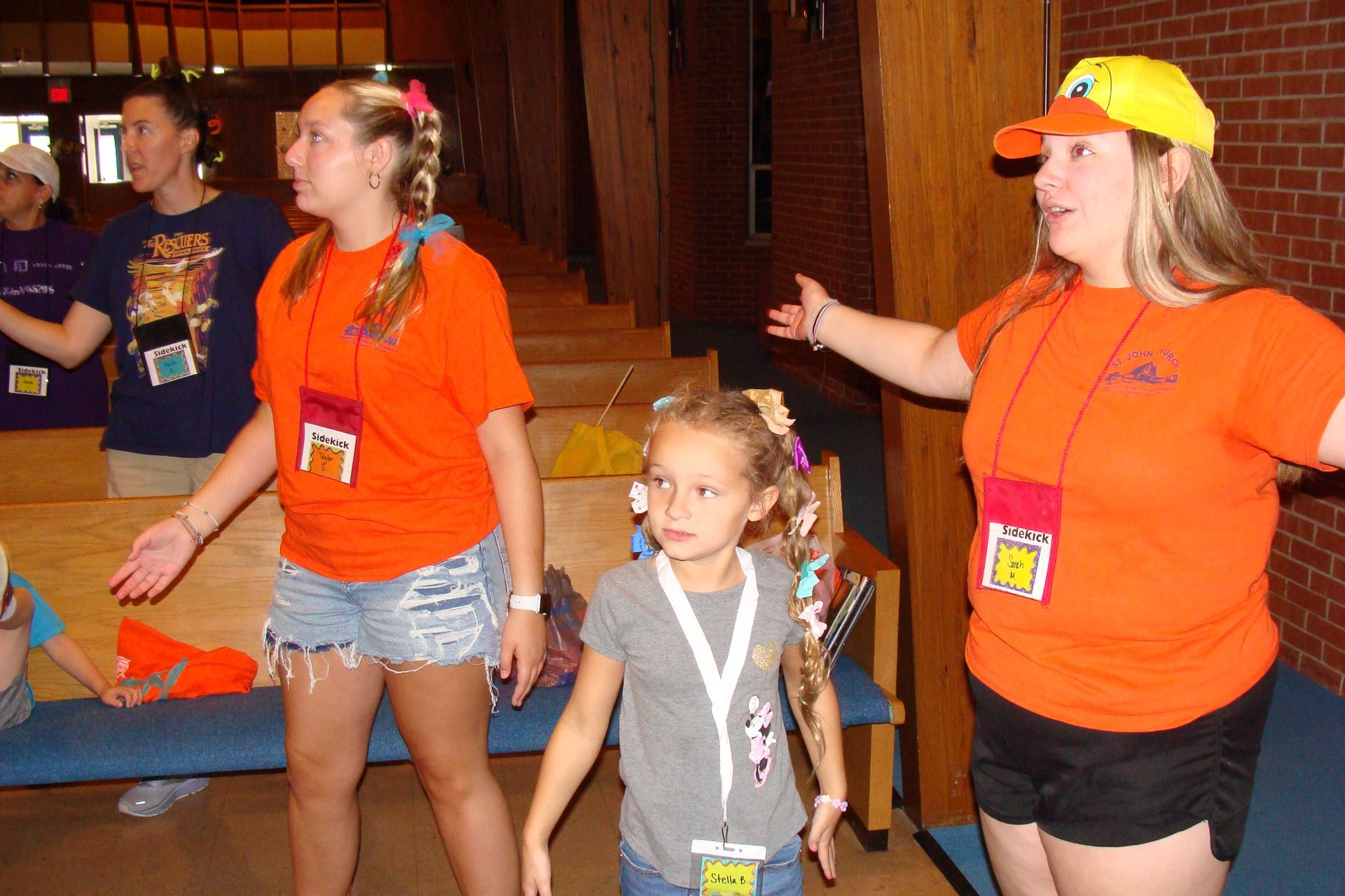 People in orange shirts and lanyards standing in a hallway, one gesturing while a child stands in front.