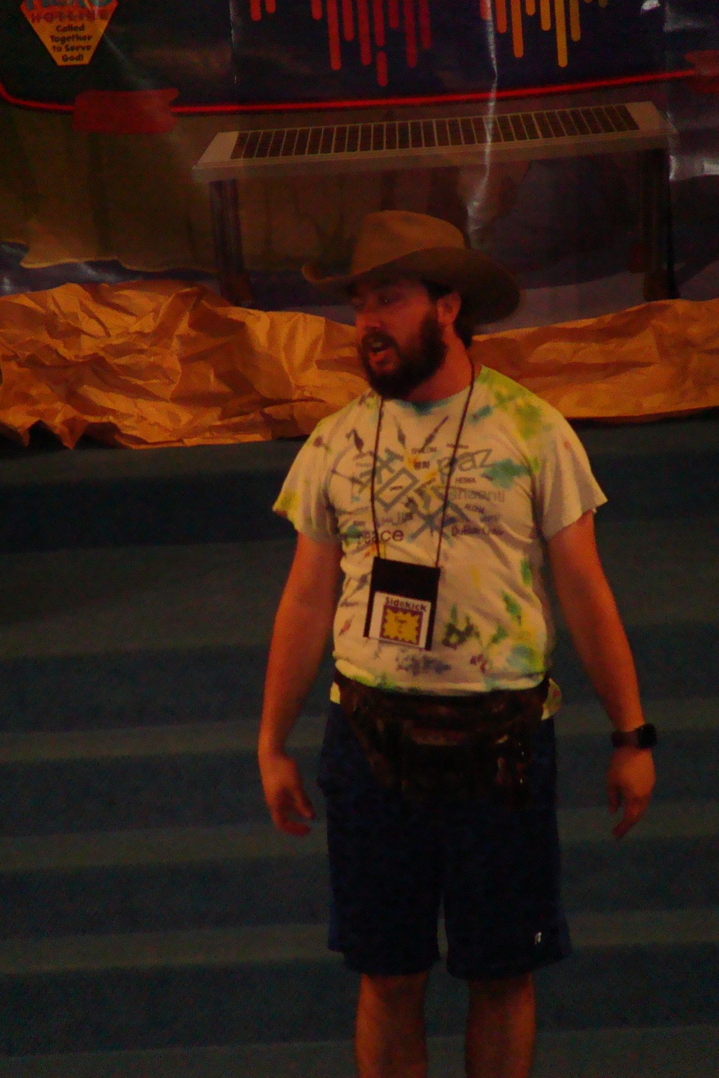 Man standing indoors in a cowboy hat, tie-dye shirt, shorts, and a lanyard, with a mural backdrop.