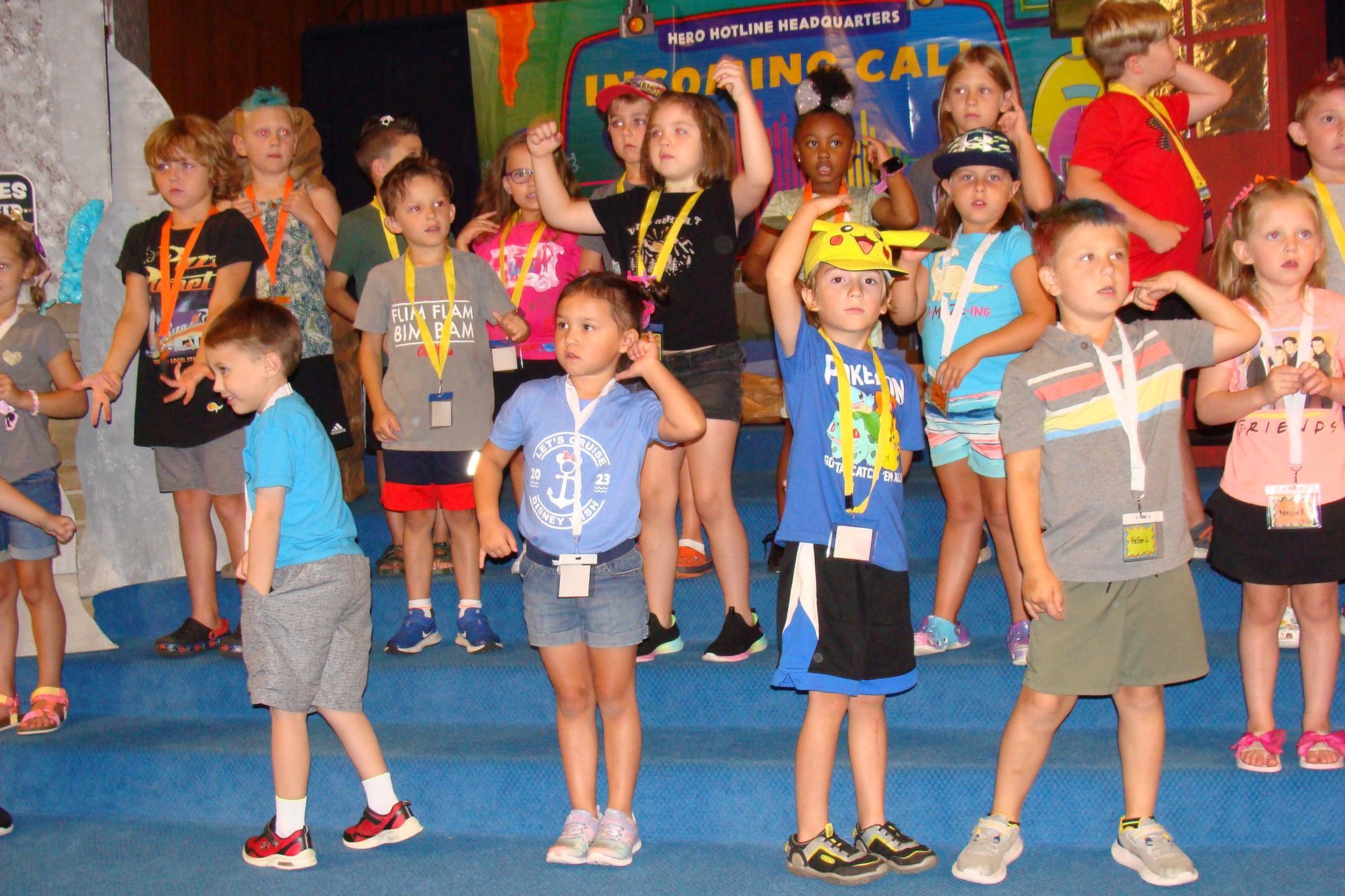 Children wearing medals and colorful hats stand on a stage during a celebration event.