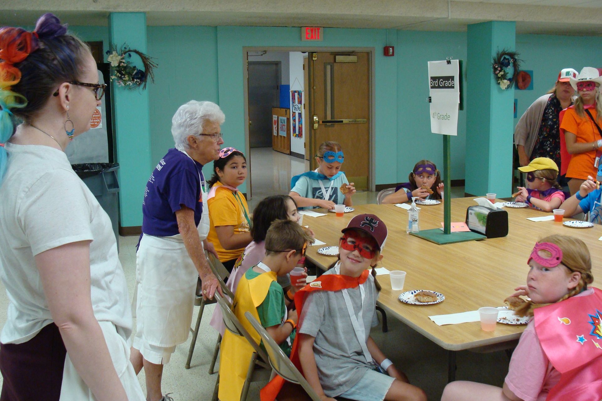Children in masks and capes sit around a table in a room while an adult stands nearby.