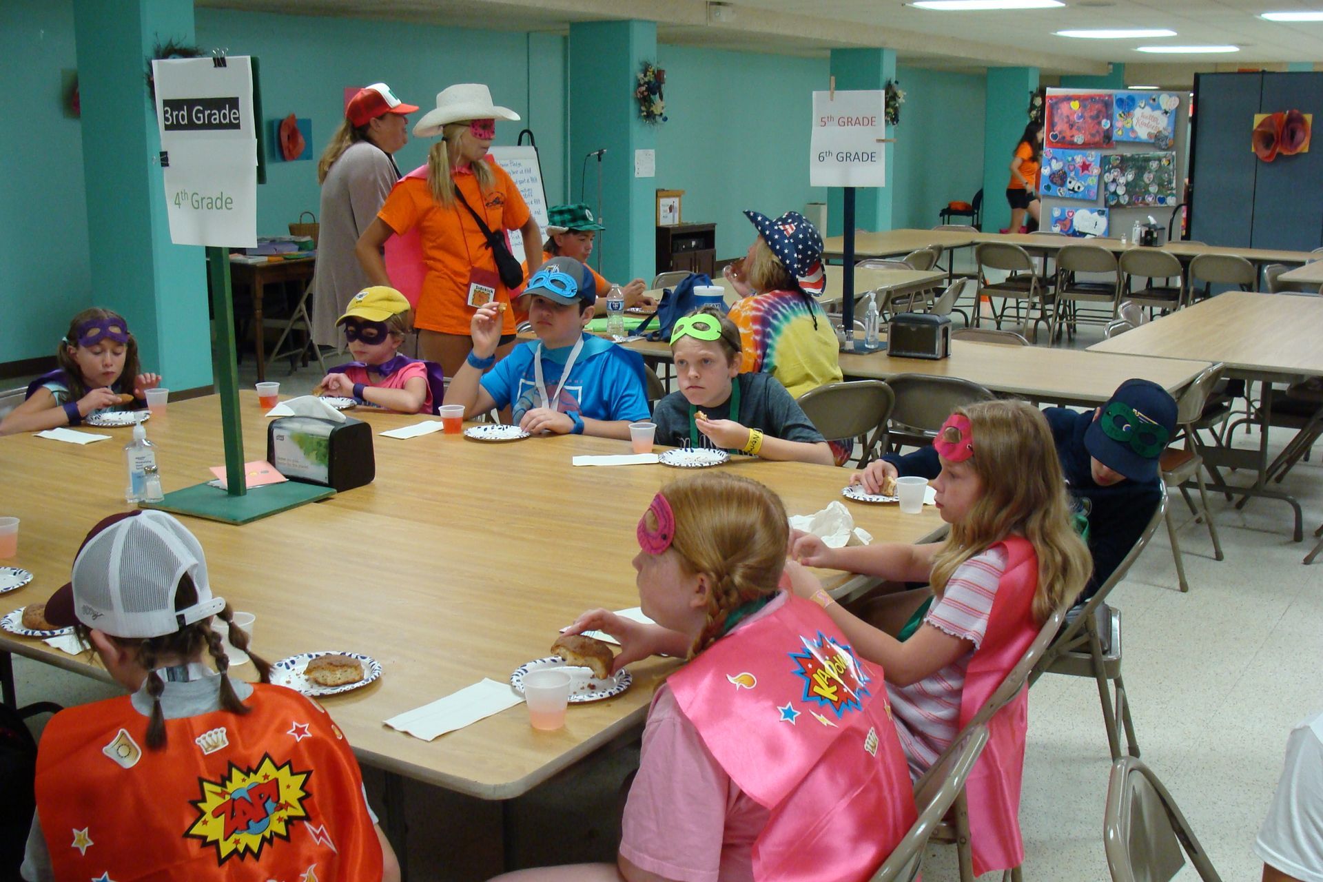 Children in costumes sit around a table at a classroom party, with an adult standing nearby.
