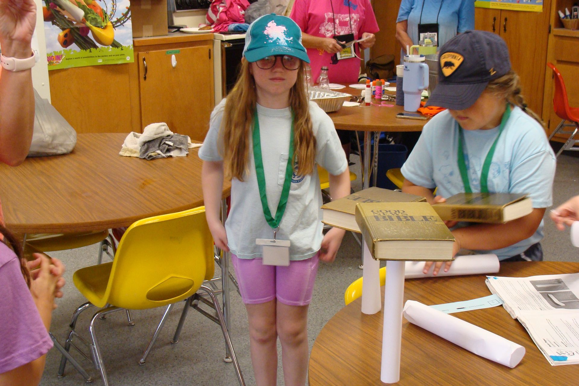 Two children with lanyards and hats stand at a table in a classroom or workshop.