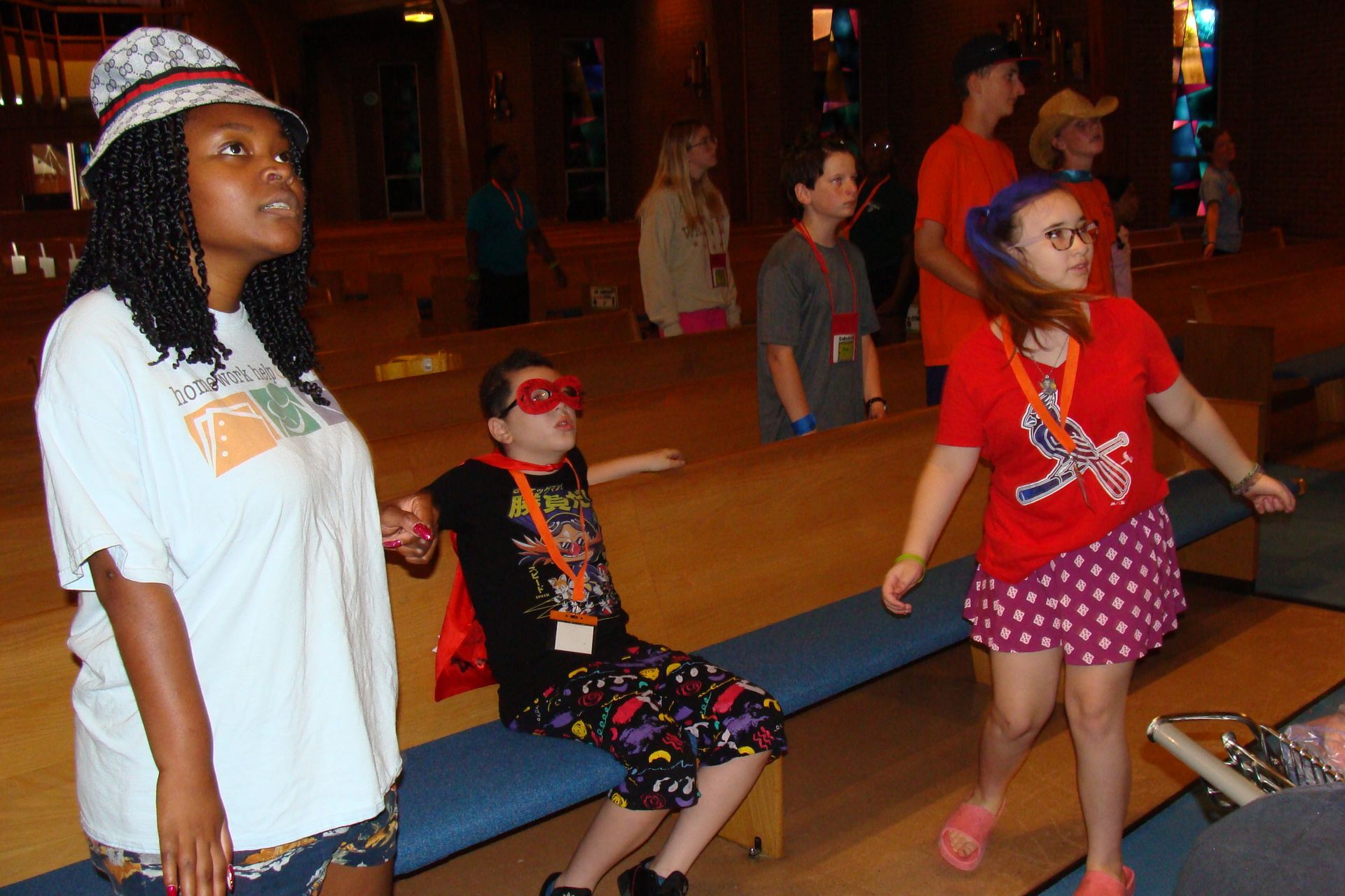 Children wearing hats and sunglasses stand and sit by a bowling lane in a dim arcade.