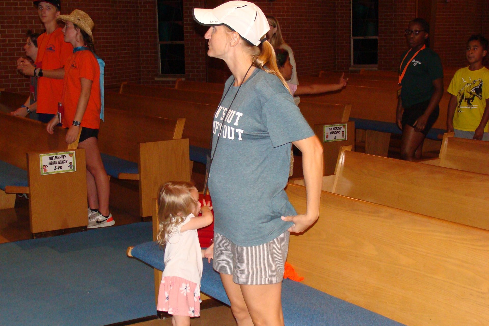 Woman in a white cap stands beside a small child in a gymnasium with wooden bleachers.