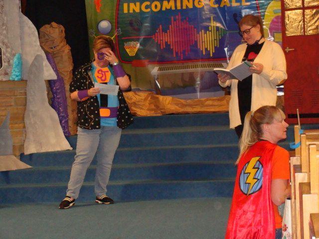Costumed people on a blue stage reading from papers at an incoming-call event sign