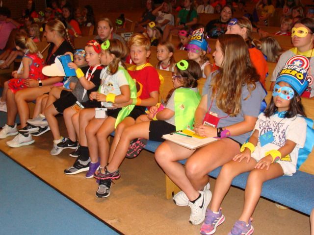 Children sitting in a gym bleacher, wearing colorful wigs and costumes at a school event
