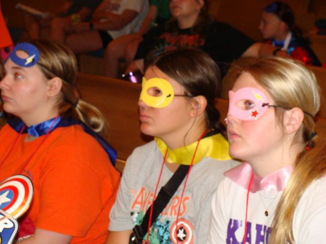 Three people in colorful face paint and costumes watching an event in a crowd