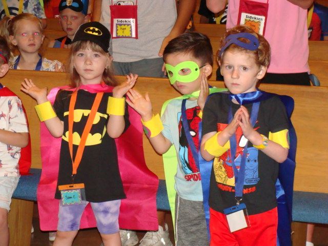 Three children in colorful superhero costumes posing with arms raised at a party
