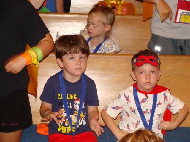 Two children seated on wooden bleachers, one in a blue shirt and one in a red mask and cape, at an event