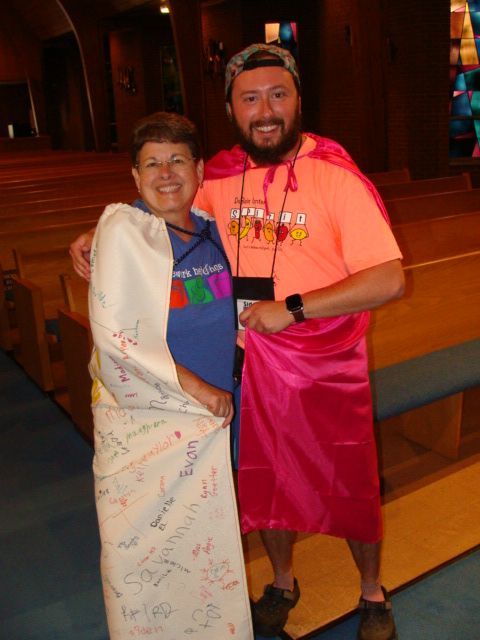 Two people smiling and posing together indoors; one wears a white wrap, the other a neon shirt and pink wrap.