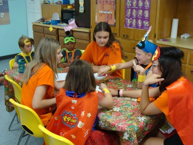 Children in orange shirts play a board game around a table in a classroom