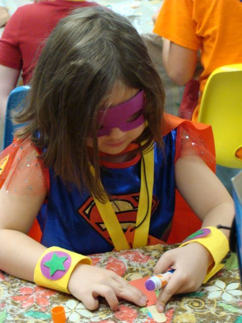Child in a superhero costume and mask coloring at a table with crayons and paper.