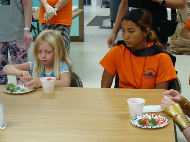 Children seated at a cafeteria table, eating from paper plates and cups