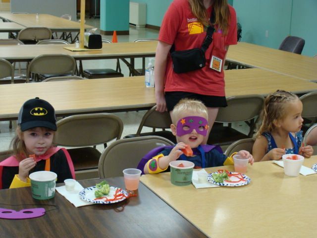 Children seated at a classroom table with snacks and cups, one wearing a purple mask, adult standing behind them