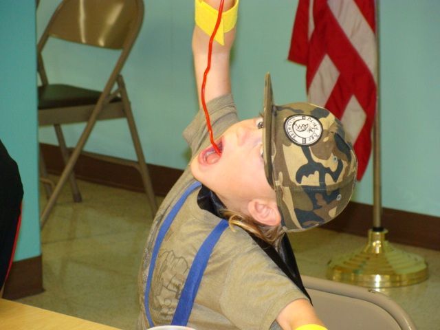 Child in camouflage cap and suspenders tilts head back to catch liquid from a bottle indoors near a flag.