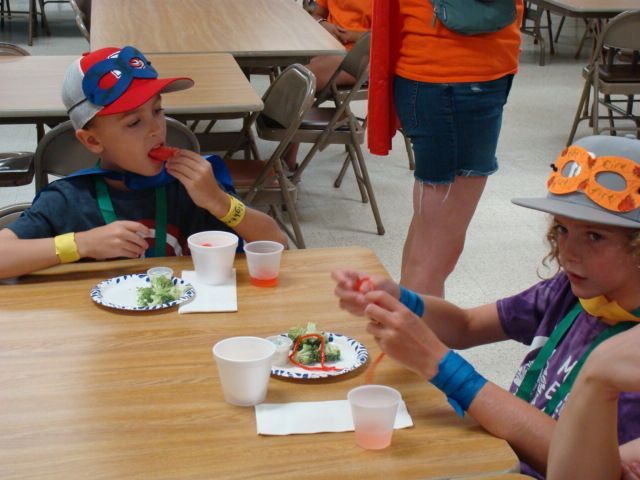 Children eating snack at a table in a cafeteria, wearing colorful hats and orange shirts.