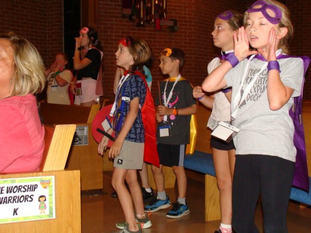 Children singing in a church choir, standing by pews with purple masks on their faces
