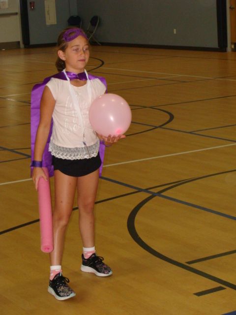 Child holding a pink balloon and bat in a gymnasium, wearing a purple costume and white top.