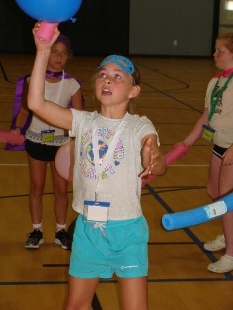 Child indoors at a gym, reaching up to catch a blue ball with a focused expression.