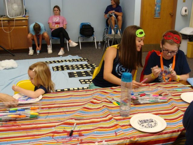 Children making crafts at a striped table in a classroom, with papers, markers, and scattered art supplies.