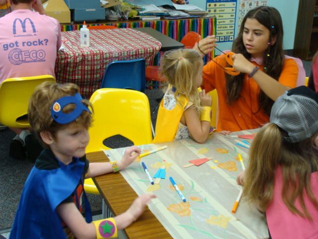 Children coloring at a table in a classroom, with one child in a blue costume and another in an orange shirt.