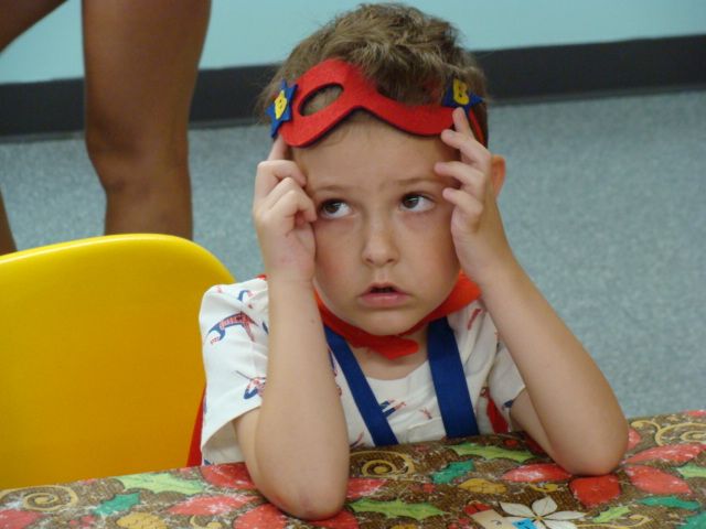 Child in a red and blue superhero mask adjusts it while sitting at a patterned table