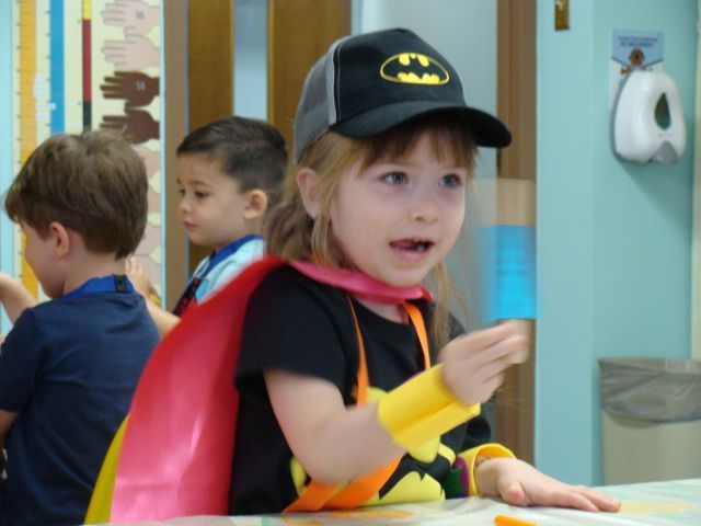 Child in Batman hat and red cape holding a brush at a classroom art table