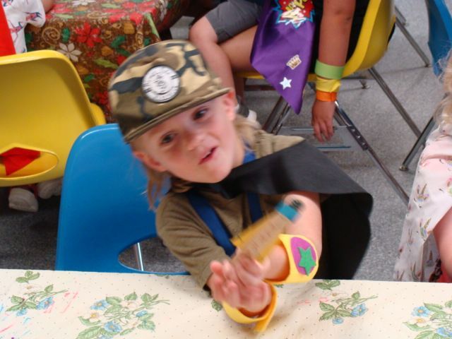 Child in a camouflage cap reaching toward the camera at a decorated table, with other children seated behind.