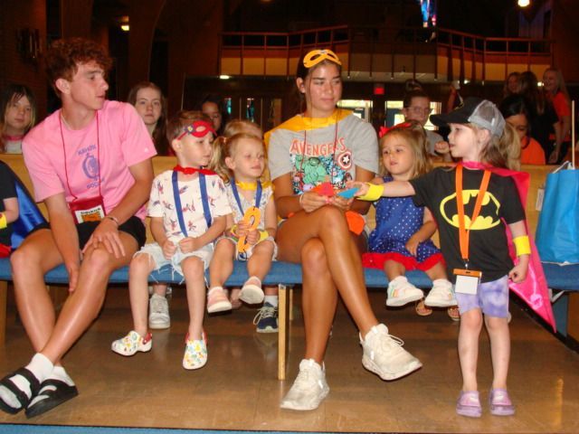 Children in colorful costumes sitting on a bench at an indoor event