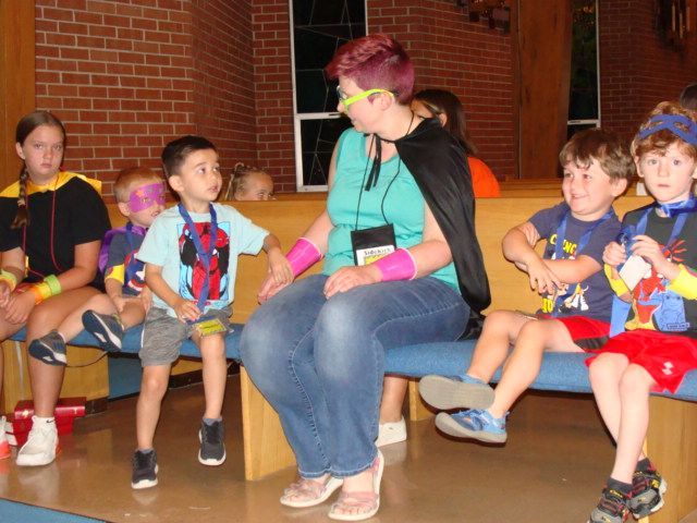 Children in colorful shirts sit on benches indoors, listening to a woman in sunglasses.