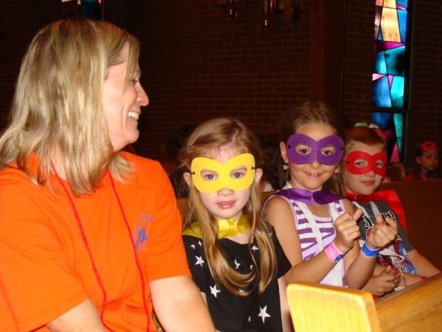 Four costumed children and an adult in orange shirts sit indoors, wearing colorful superhero masks.