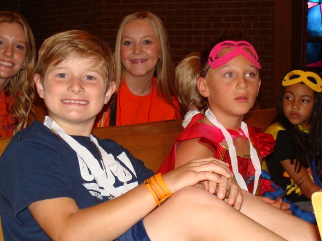 Children seated together indoors, smiling at the camera, some wearing bright headbands and costumes