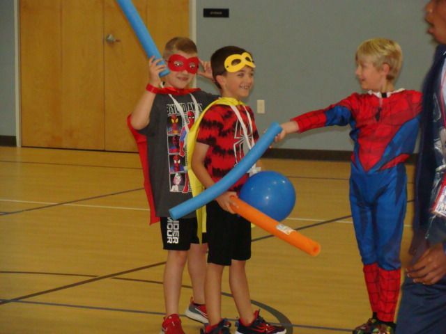 Three children in superhero costumes holding balloon swords in a gymnasium