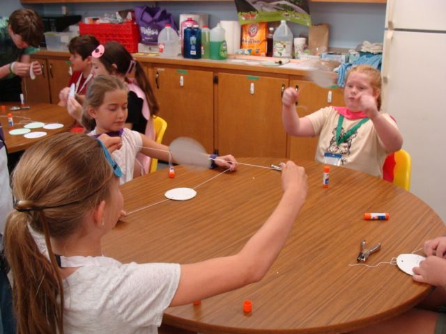 Children doing a craft activity around a round table in a classroom, with supplies scattered on the table.