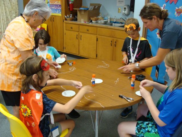 People seated around a table in a classroom, painting crafts and talking with an instructor nearby