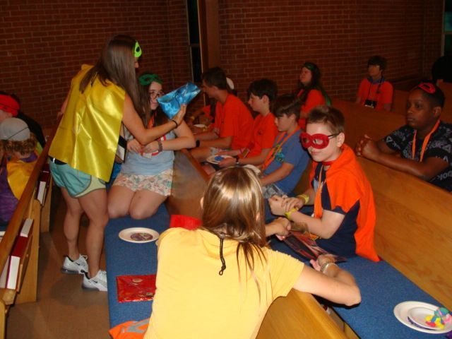 Children in colorful costumes sit around tables at a party or classroom activity in a brick-walled room.