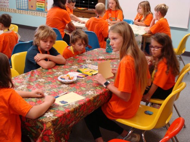 Children in orange shirts sit around a table with snacks and crafts in a classroom.
