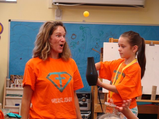 Two people in orange shirts balance a yellow ball above a black vase in a classroom.