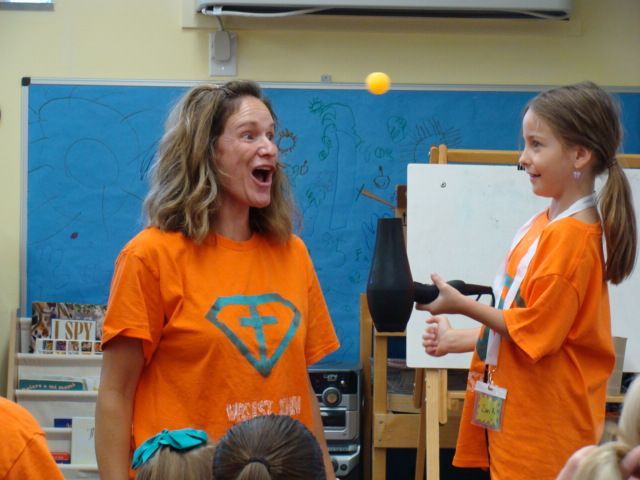 Two people in orange shirts smile at a floating ball in a classroom.
