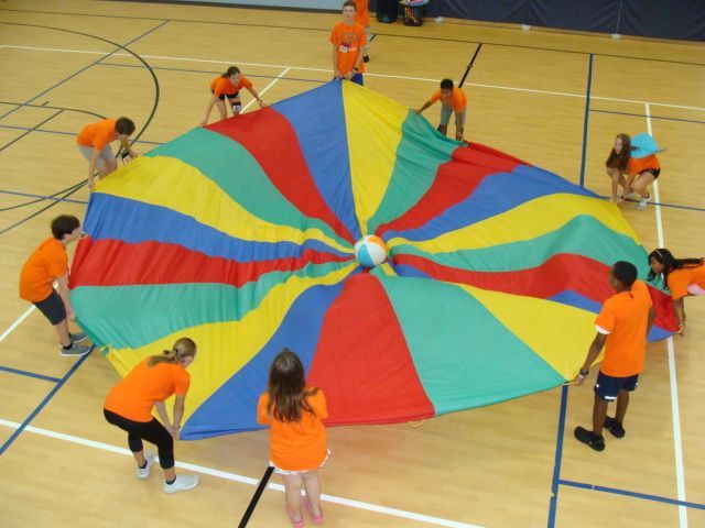 Children holding a colorful parachute in a gymnasium, forming a circle around it.