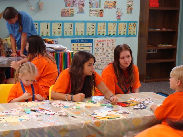 Children in orange shirts doing arts and crafts at a classroom table with an adult nearby