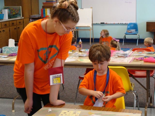 Two children in orange shirts working on a craft project at a classroom table