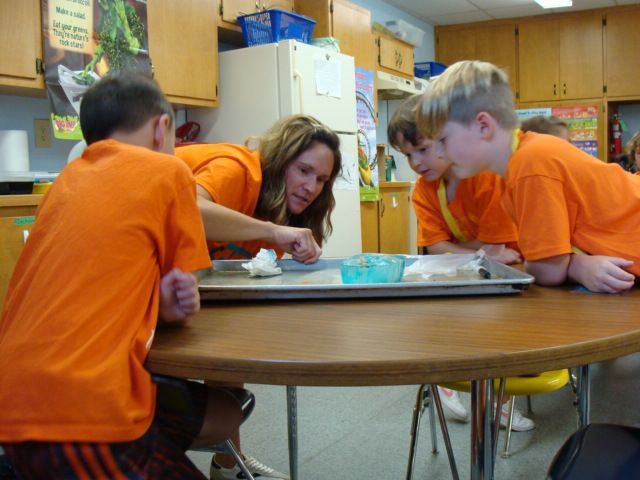 Children in orange shirts lean over a table, working together on a tray in a classroom.
