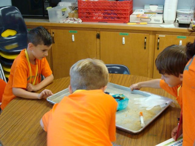 Children in orange shirts gather around a table with a tray of materials in a classroom.