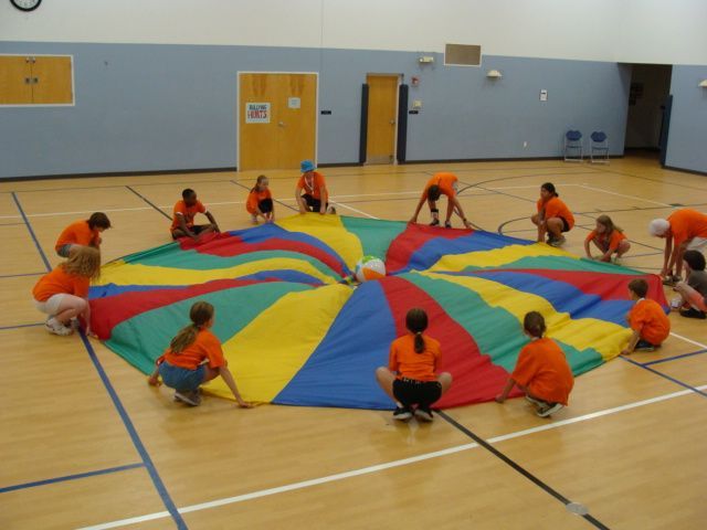 Children in orange shirts spread a colorful parachute in a gymnasium.