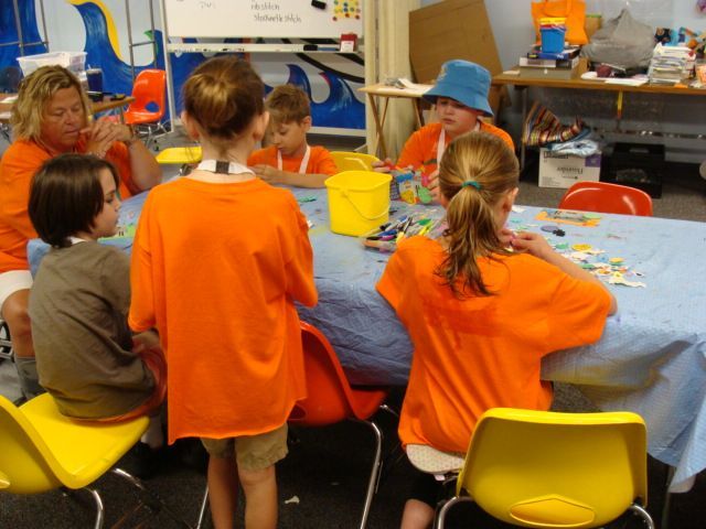 Children seated around a table in a classroom, doing a group art or craft activity with colorful supplies.